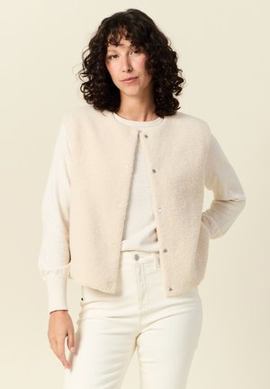 Lichtbeige fleece vest met drukknoopsluiting aan de voorkant en lange mouwen, gedragen over een crèmekleurig shirt en in combinatie met witte broek op een neutrale achtergrond.