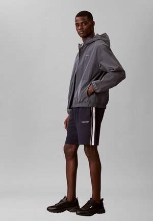 Homme mannequin debout, portant une veste à capuche grise, un short Calvin Klein noir avec des bandes blanches, et des chaussures de sport noires sur un fond gris.