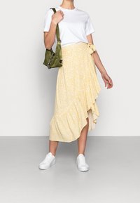 Gonna midi gialla a fiori con orlo asimmetrico, dettagli arricciati, abbinata a una t-shirt bianca e una borsa a spalla verde. Sneakers bianche completano l'outfit.