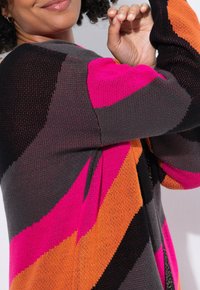 Personne portant un pull en tricot multicolore avec des rayures diagonales roses, orange, noires et grises, levant son bras près de son visage.