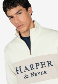 Sudadera de medio cierre en forro polar color blanco roto con una banda beige que presenta letras en azul marino "HARPER & NEYER." Cierre metálico, textura suave.