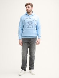 Lichtblauwe hoodie met trekkoorden en bedrukte tekst. Gepaarde met grijze jeans en witte sneakers. Soepele textuur en casual pasvorm.