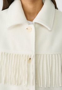 Giacca bianca in misto lana con colletto ampio, caratterizzata da due grandi bottoni e dettaglio frangiato nella parte inferiore per texture e contrasto di design.