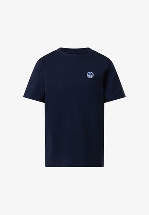 Garçon debout, mains dans les poches, portant un t-shirt bleu marine avec un petit logo de boussole et un pantalon de jogging assorti, ainsi que des baskets blanches, sur un fond gris.
