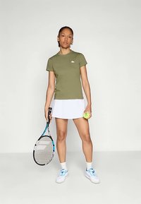 Olivengrønn tettsittende t-skjorte med en liten logo, sammen med et hvitt tennis skjørt. Holder en tennisracket og en knallgul ball.