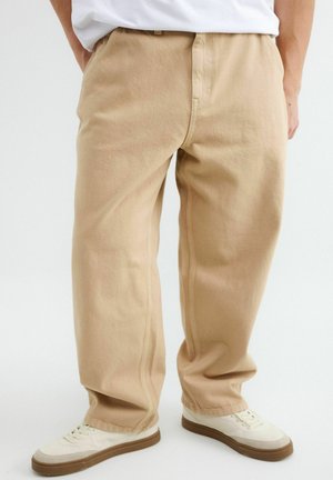 Pantalon beige à jambes larges porté avec des baskets blanches à semelles en gomme, associé à un t-shirt blanc rentré, montré de la taille aux pieds.