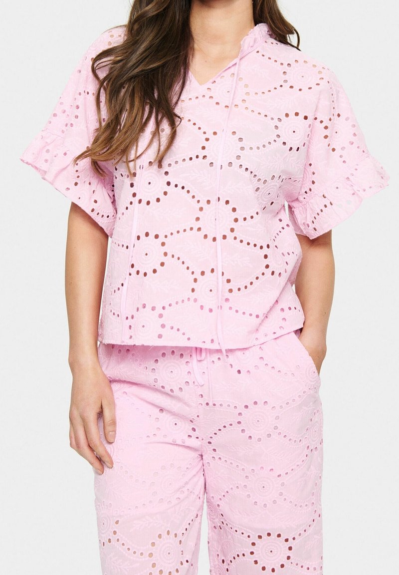 Femme portant un haut en tissu œillet rose clair assorti avec des manches courtes à volants et un pantalon avec poches, debout devant un fond uni.