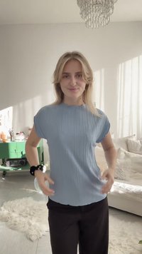 Μια νεαρή γυναίκα με ξανθά μαλλιά φοράει ένα ανοιχτό μπλε πλισέ τοπ και μαύρο παντελόνι, στέκεται σε ένα φωτεινό σαλόνι με λευκά και πράσινα έπιπλα.