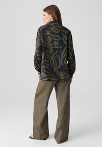Blusa blu con grandi motivi a foglia verde oliva, maniche lunghe e polsini con bottoni, abbinata a pantaloni larghi in lino beige chiaro.