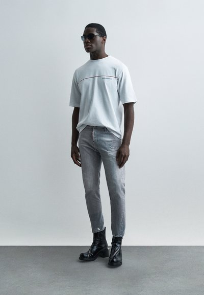 Homme portant des lunettes de soleil noires, un t-shirt blanc oversize DSQUARED2, un jean gris délavé déchiré et des bottines en cuir noir, debout contre un mur uni.