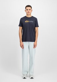 Σκούρο μπλε κοντομάνικο T-shirt με χρυσό λογότυπο στο στήθος, συνδυασμένο με ανοιχτό μπλε φαρδιά τζιν και γκρίζα αθλ�ητικά παπούτσια.