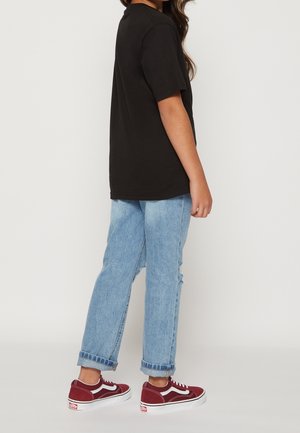 Person står sidelæns iført en sort t-shirt, lyseblå op rullede jeans og røde lave sneakers med hvide såler.