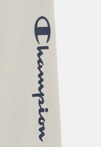 Lys beige stoff med en mørk marineblå "Champion"-logo i kursiv. Designet er enkelt med en jevn tekstur og uten tilleggsmønstre.