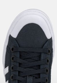 Sneaker in tela grigio scuro con punta in gomma bianca, lacci neri piatti e occhielli metallici; presenta dettagli laterali in bianco.