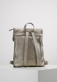 Metallinen hopeansävyinen nahkareppu, jossa on säädettävät hihnat, yläkahva ja pystysuora sivuvetoketjupussi, asetettuna valkoiselle jalustalle.