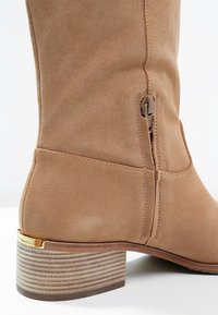 Botte haute jusqu'au genou en suédine beige avec une fermeture éclair latérale, un talon empilé et un accent doré à la base du talon ; texture lisse et coutures nettes.