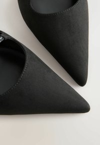 Ballerines noires en daim à bout pointu avec doublure intérieure lisse, photographiées sur un fond neutre clair.