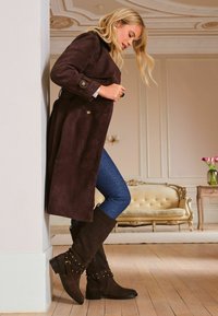Hnědý semišový kabát s páskem v pase, v kombinaci s tmavě modrými džíny a hnědými kozačkami po kolena, které mají dekorativní nýty a přezky.