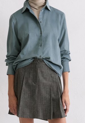 Personne portant une chemise bleue boutonnée sur un col roulé beige et une jupe plissée grise, debout contre un mur blanc uni.