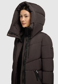 Pruun pufjakker suurte kapuutsiga, millel on teibitud tekstuur, lukugaTaskud ja reguleeritavad nöörid. Lukuga esiotsa sulgus.