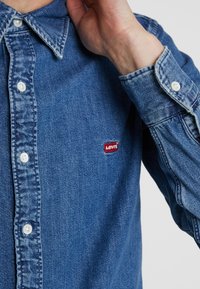 Spijkershirt in medium blauw, met een button-down kraag, witte knopen en een klein rood Levi's-logo op het linker borstzakje.