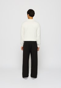 Mand stående med ryggen til, iført en hvid langærmet skjorte, sorte bukser med vide ben og hvide sneakers på en neutral baggrund.