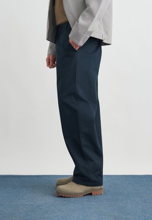 Persona in piedi su un tappeto blu che indossa pantaloni blu navy, giacca grigio chiaro e stivali beige in camoscio.