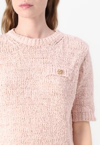 Pull en laine rose clair avec un col rond, des manches courtes et une poche poitrine ornée d'un bouton doré. La texture est lâche et volumineuse.