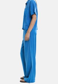 Osoba ubrana w luźne niebieskie spodnie i koszulę z krótkim rękawem, stoi bokiem z gołymi stopami w czarnych klapkach na tle jednolitym.