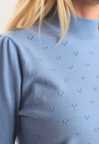 Ljust blått stickat tröja med hög ribbad krage, med små perforerade mönster över hela och lätt puffade ärmar.