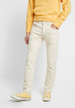 Personne portant un jean couleur crème, un sweat-shirt jaune et des baskets jaunes assorties, debout devant un fond blanc uni.