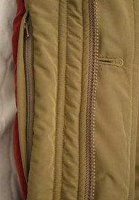 Puderet jakke med grønt ydre, med en brun lynlås, ribbet struktur og kontrasterende rødt stof, der titter frem fra siden.