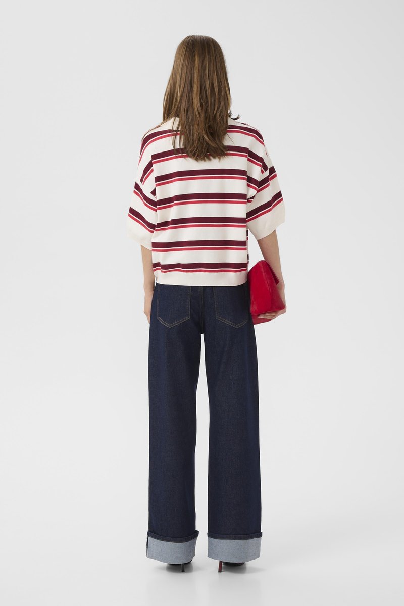 Woman with long hair wearing a white and red striped short-sleeve top, wide dark blue jeans cuffed at ankles, holding a red clutch.
