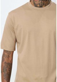 Chemise beige à manches courtes en tissu lisse et extensible. Présente un col rond et une coupe décontractée, avec des tatouages visibles sur le bras.