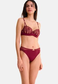 Ensemble de lingerie bordeaux comprenant un soutien-gorge en dentelle transparente avec des motifs floraux et un brief assorti avec des accents en dentelle et un détail en métal doré.