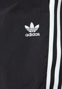 Sorte treningsbukser med hvite tre-stripes accentuerte detaljer på sidene. Har et brodert hvitt Adidas-logo på venstre lår.