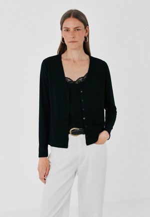Femme portant un cardigan noir sur un haut en dentelle noire et un pantalon blanc, debout avec une main dans la poche devant un fond uni.