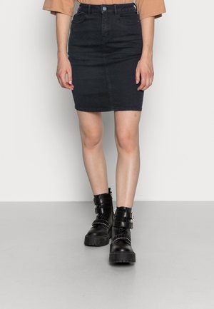 Femme portant une jupe en jean noire et des bottines en cuir noires avec des boucles, debout sur un sol gris contre un mur blanc.