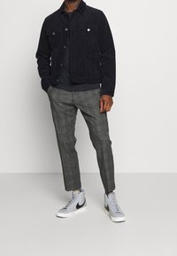 Man som bär en svart kordflorsjacka, svart skjorta, grå rutiga capribyxor och grå höga sneakers med svarta swoosh-loggor.