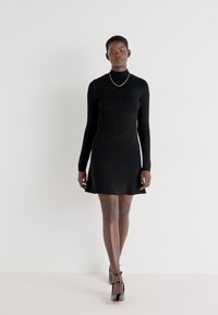 Fekete, hosszú ujjú ruha, testhezálló fazonnal és ellapult szegéllyel. Sima kötött textúra, minimalista dizájn, fekete magassarkú cipővel és egy egyszerű nyaklánccal párosítva.