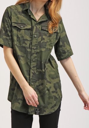 Chemise à manches courtes en camouflage vert avec un devant à boutons, deux poches poitrine et un ourlet arrondi, confectionnée en tissu de coton léger.