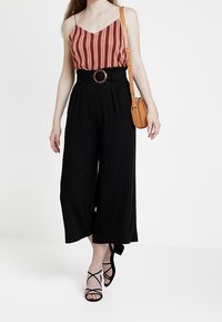 Red striped sleeveless top paired with high-waisted black wide-leg trousers, accented by a brown circular belt and strappy black heels.