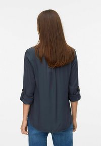 Blouse bleu marine foncé avec une texture lisse, manches longues retroussées et détails de boutons. L'arrière présente un ourlet arrondi. Associée à un jean bleu.