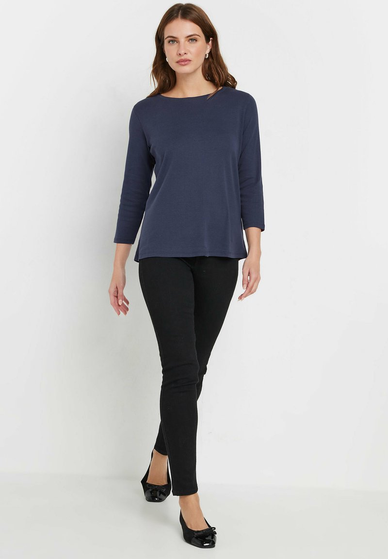 Navyblaues Langarmoberteil mit weitem Rundhalsausschnitt, kombiniert mit schwarzen Skinny-Jeans und schwarzen patentierten Ballerinas mit einer Schleifendetail. Einfaches und figurbetontes Design.