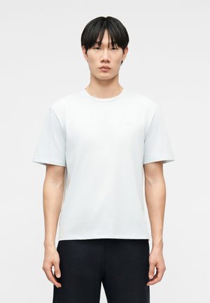 Bărbat tânăr cu păr negru, stând cu fața spre înainte, purtând un tricou alb simplu cu mânecă scurtă și pantaloni negri, pe un fundal alb.