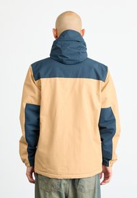 Man wearing beige and dark blue hooded jacket, viewed from behind, with light denim jeans against a plain white background.