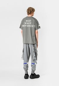 Camiseta de algodón gris con la impresión "MOVIMIENTO MOD WAVE" en la parte posterior, combinada con pantalones cargo que tienen múltiples bolsillos y acentos grises. Calzado negro.