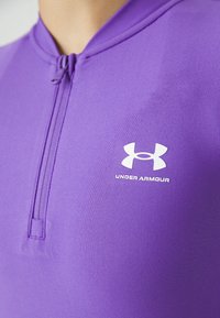 Lilla treningstop med halv-glidelås, laget av glatt, stretch-stoff. Under Armour-logoen er vist i hvitt på brystet.