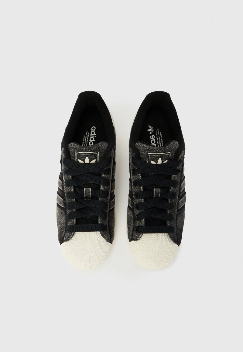 Paire de baskets Adidas noires avec bouts renforcés en caoutchouc blanc et lacets noirs, vues de dessus sur un fond blanc.