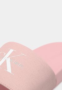 Rózsaszín Calvin Klein papucs, texturált pánttal és nagy fehér "CK" logóval, sima fehér háttérrel.